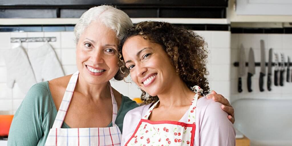 A senior mother and her daughter are smiling and standing in the kitchen together.