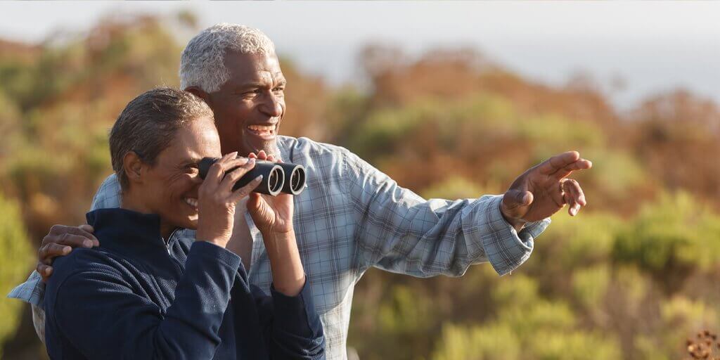 A senior couple outside pointing in the distance and looking through binoculars.