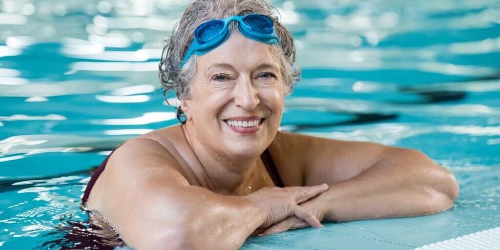 Elderly woman in pool Mature woman wearing swim goggles at swimming pool. Fit active senior woman enjoying retirement standing in swimming pool and looking at camera. Happy senior healthy old woman enjoying active lifestyle.