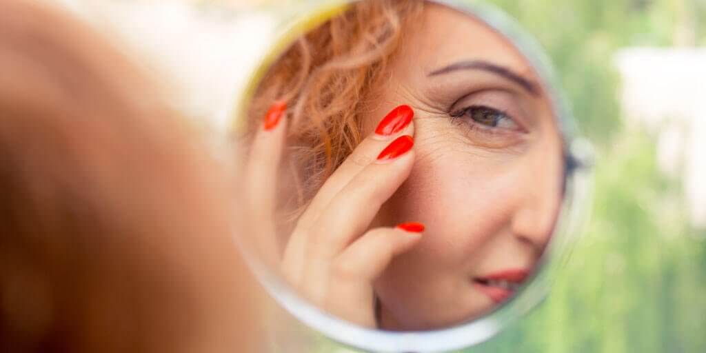 what-is-oculoplastic-cosmetic-surgery-1024x512 A middle-aged woman is touching her face while looking in a mirror.