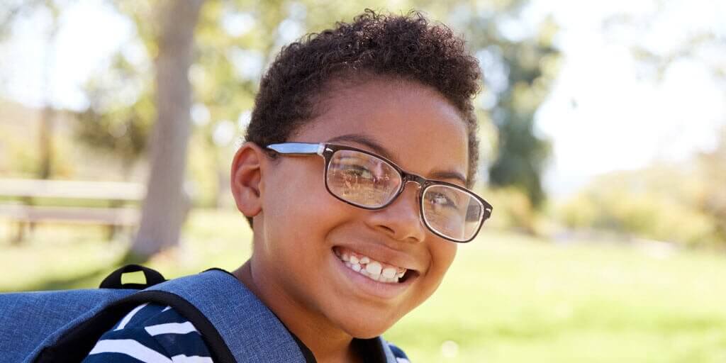 A young boy with eyeglasses and a backpack is smiling outdoors.